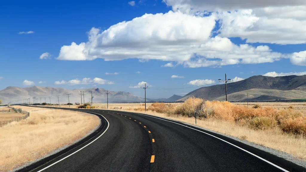 Open highway curving through golden grassland toward distant mountains under a big blue sky
