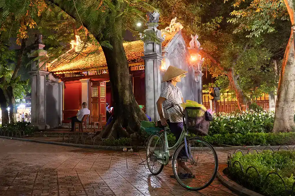 Woman with a bicycle beside an illuminated temple gate at night in Hanoi, Vietnam