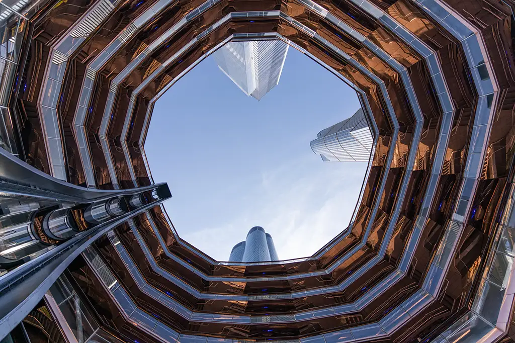 Looking straight up through the spiraling honeycomb staircases of The Vessel in New York City, framed against the sky and surrounding skyscrapers